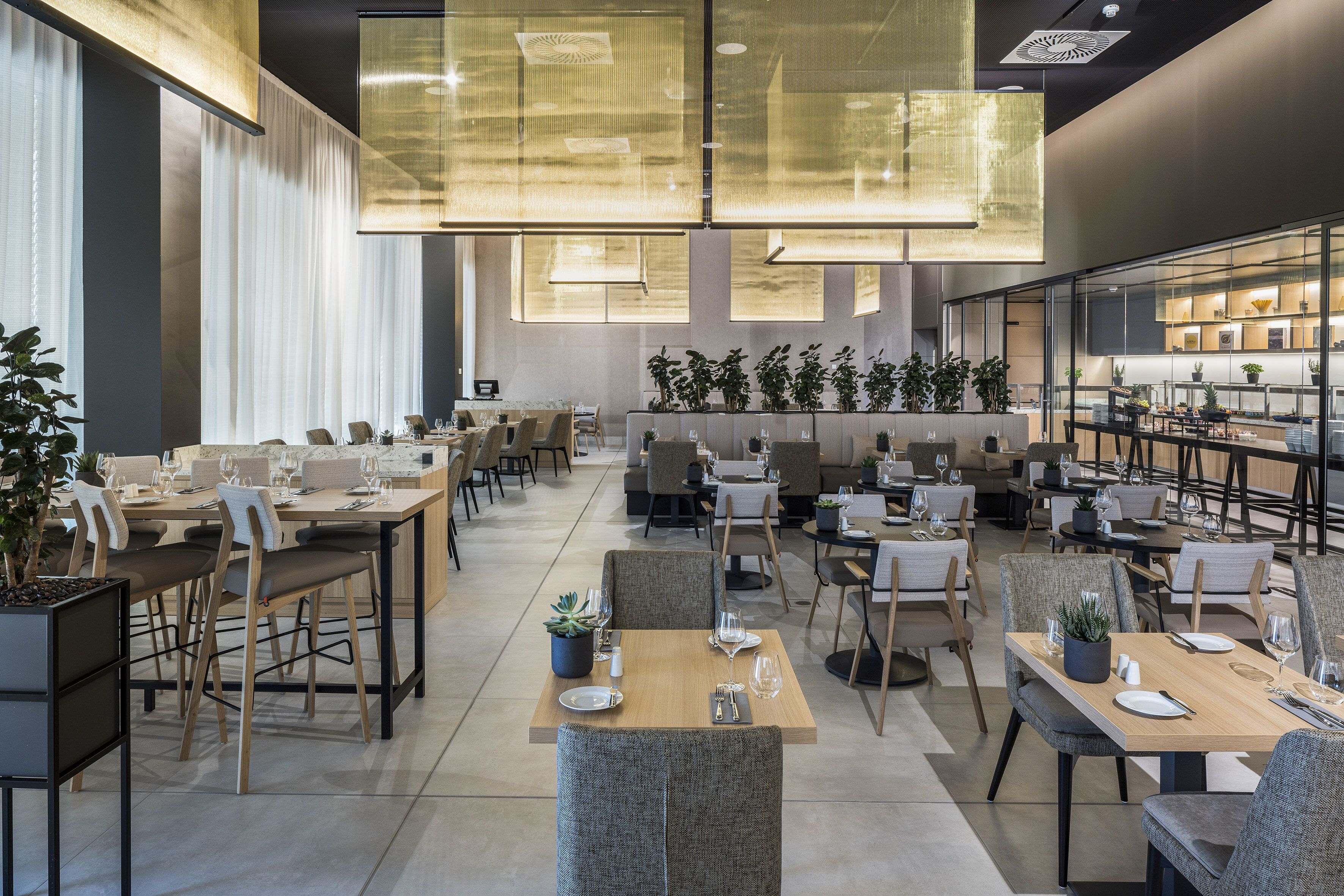A modern dining area with multiple tables and chairs, large hanging lights, and potted plants.