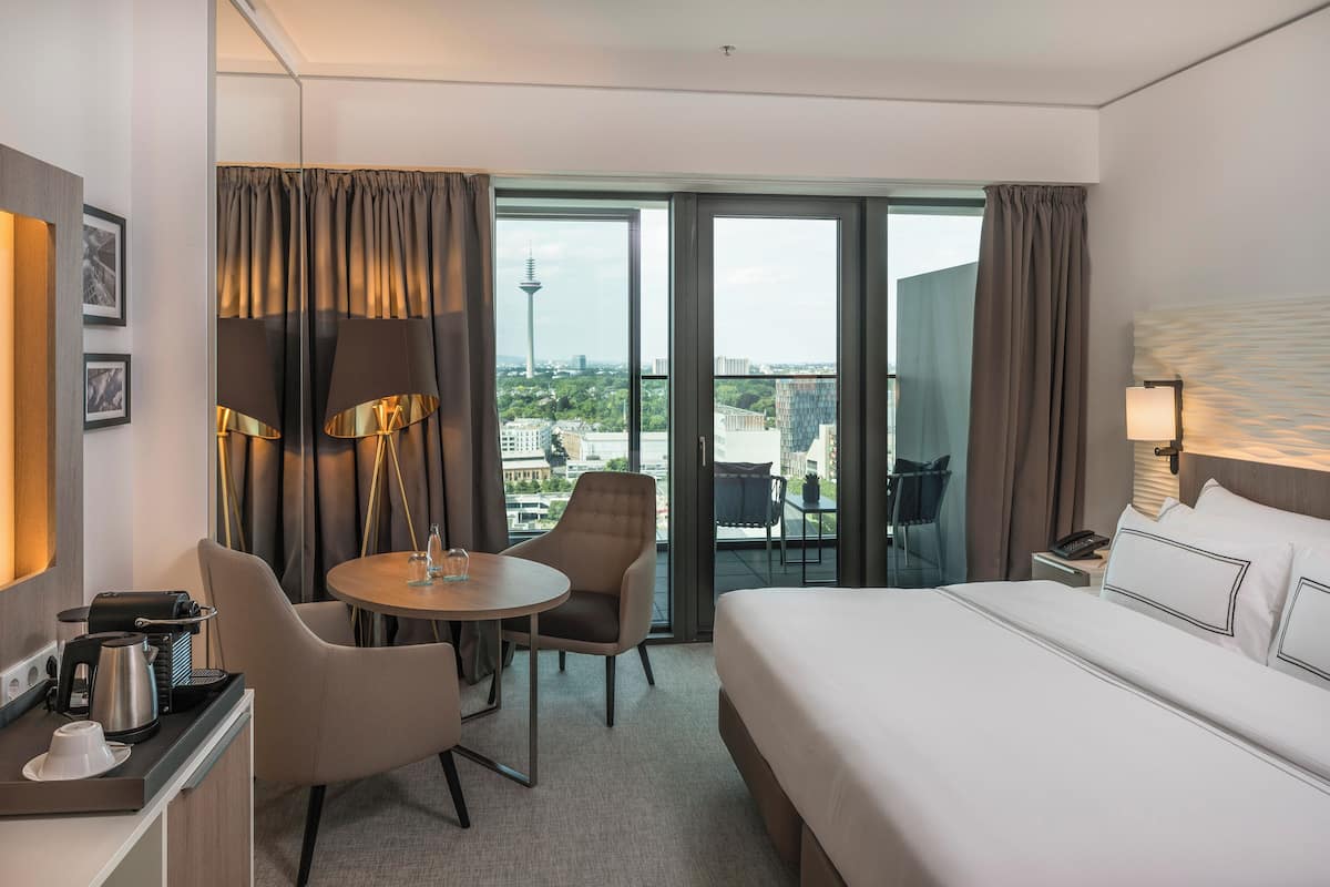 Ein Hotelzimmer mit Bett, Schreibtisch mit Kaffeemaschine, Stuhl, Tisch und Blick auf die Stadt durch große Fenster.