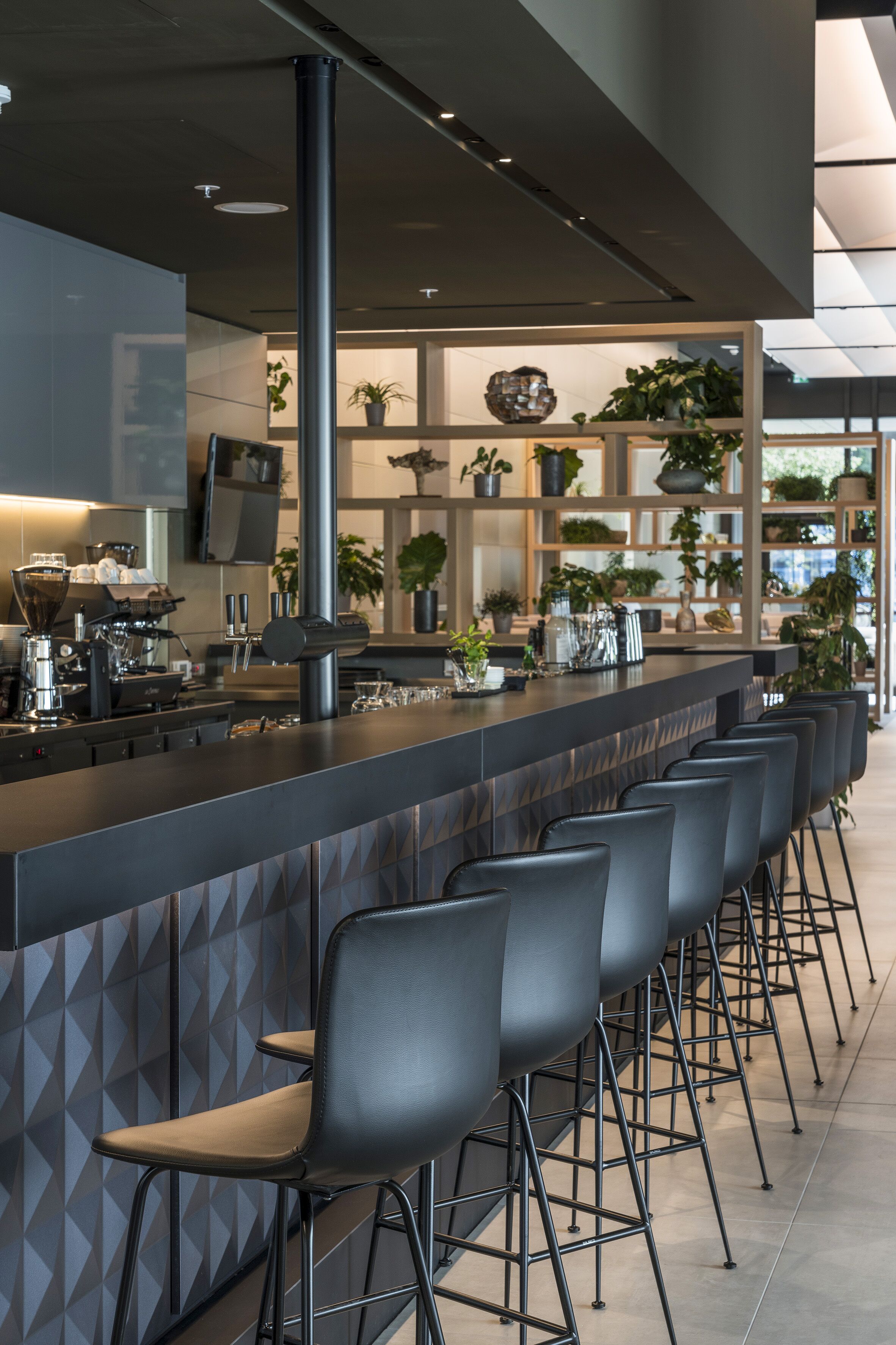 A modern bar area with a long counter, multiple stools, and a variety of plants.