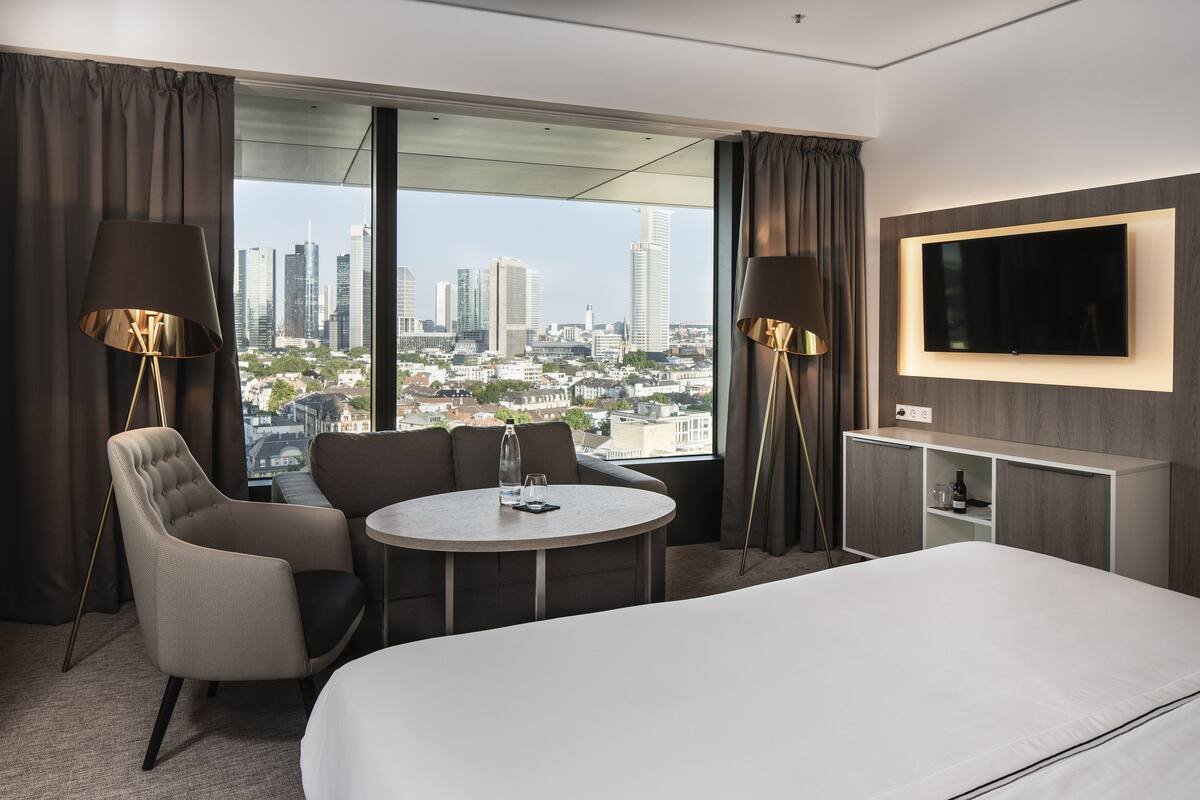 Ein Hotelzimmer mit einem großen Bett, einem runden Tisch, zwei Sesseln, einem an der Wand befestigten Fernseher und Blick auf eine Stadtlandschaft durch große Fenster.