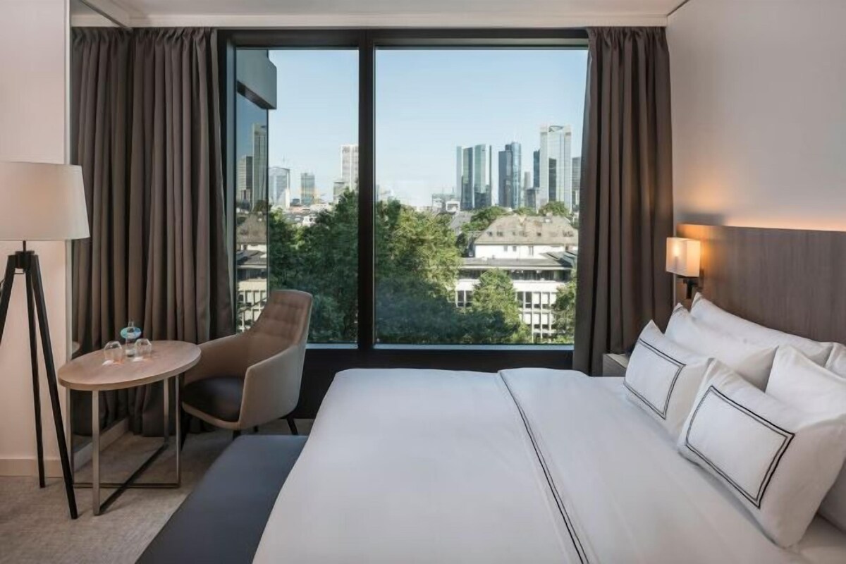 A hotel room with a large bed, a chair, a small table, and a view of the cityscape through large windows.