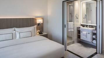 Une chambre d’hôtel moderne avec un lit soigneusement fait, une table de chevet avec une lampe et une salle de bain visible à travers une porte en verre.