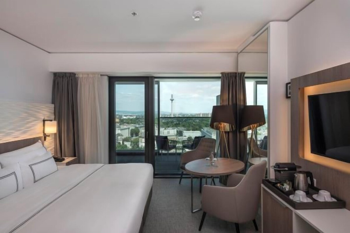 A hotel room with a bed, a desk with a chair, a TV, and a view of the cityscape.