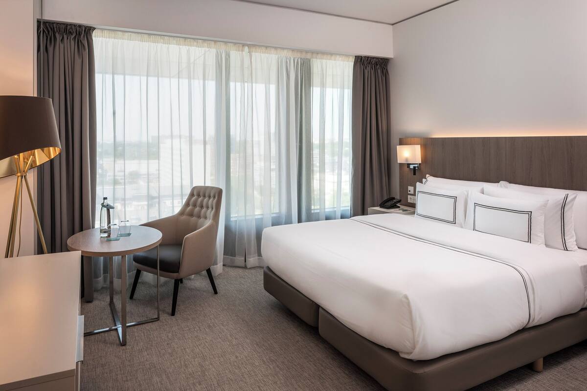 Ein Hotelzimmer mit einem großen Bett, einem runden Tisch, einem Sessel und Blick auf die Stadt durch transparente Vorhänge.