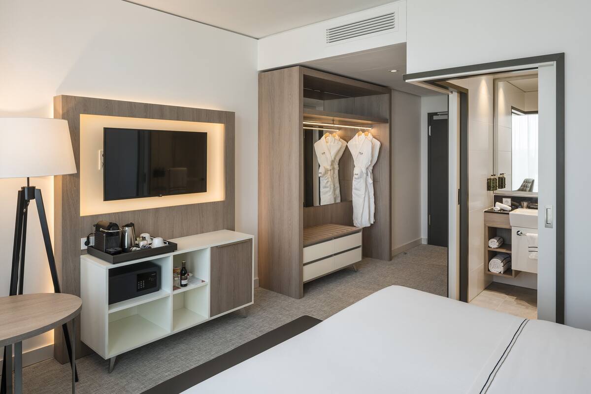 Ein modernes Hotelzimmer mit einem großen Bett, einem Nachttisch mit Lampe, einem an der Wand montierten Fernseher, einem begehbaren Kleiderschrank mit Bademänteln sowie einem Badezimmer mit Spiegel und Ablagen.