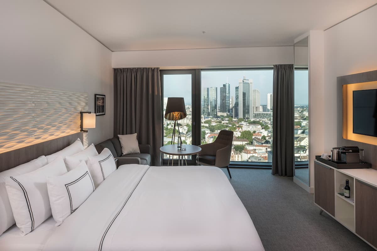 Ein Hotelzimmer mit einem großen Bett, einem Schreibtisch, einem Sessel, einem Fernseher und Blick auf die Stadt.