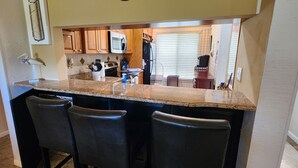 Una cocina con encimeras de granito, armarios de madera, microondas y refrigerador. Hay tres taburetes de bar y una ventana con persianas.