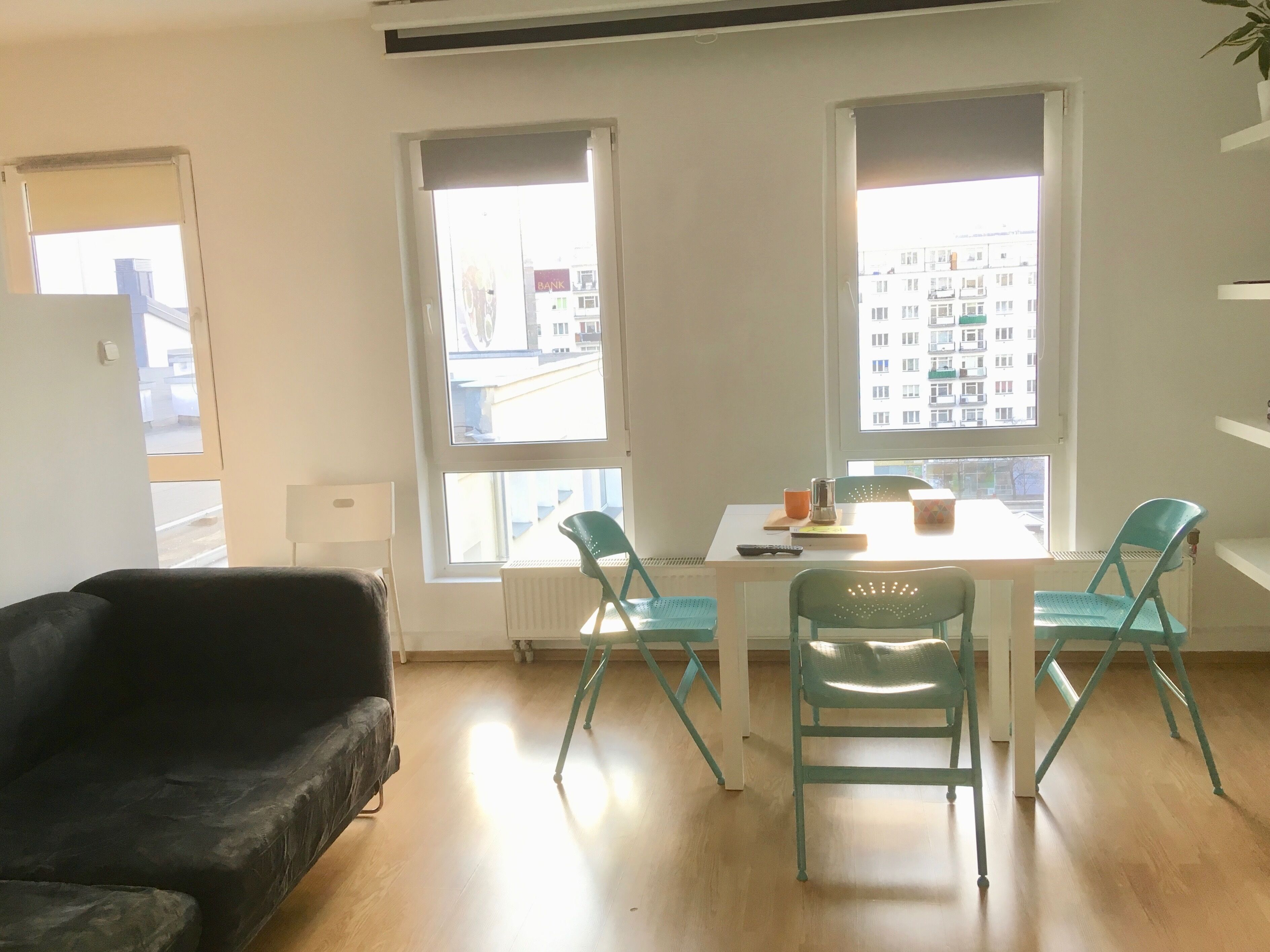 A room with a black sofa, two teal chairs, a white table, and a window with a view of buildings.