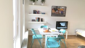 Una habitación con una mesa blanca, sillas color esmeralda, una estantería, un televisor y una cama con cubierta estampada.