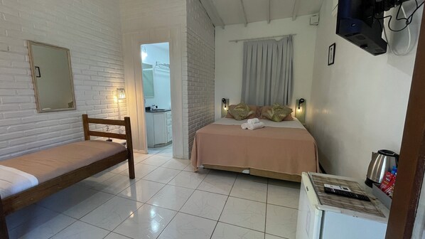 A bedroom with a bed, a chair, a small table with a kettle, and a bathroom visible in the background.