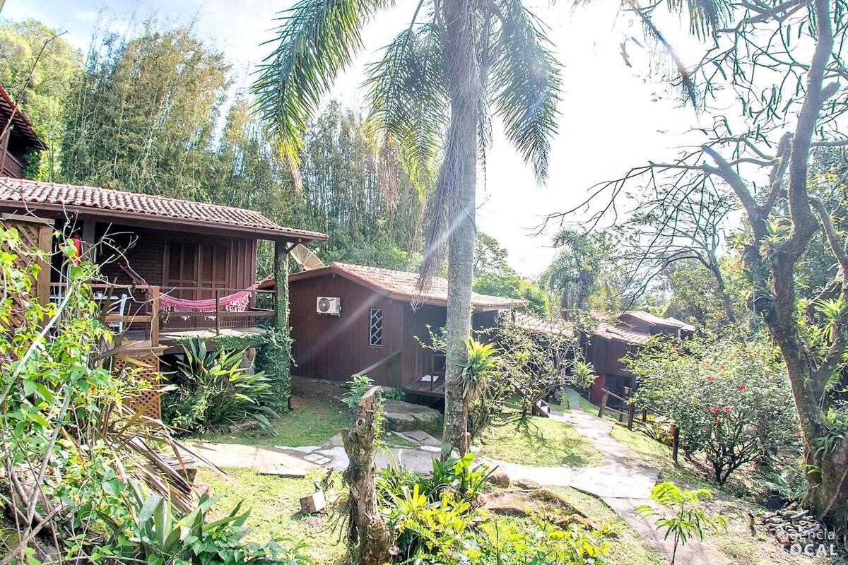 A rustic wooden house with a tiled roof, surrounded by lush greenery and palm trees.