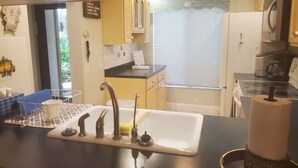 A compact kitchen with a sink, faucet, and countertop. There is a window with blinds, a refrigerator, and a coffee maker.