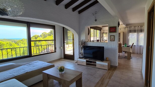 A spacious living area with a large TV, wooden furniture, and a balcony with a scenic view. - Detached house with terrace in the middle of a pine forest with sea view (Altea la vella)