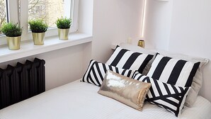 A bedroom with a bed, black and white striped pillows, a gold throw pillow, and three potted plants on the windowsill.