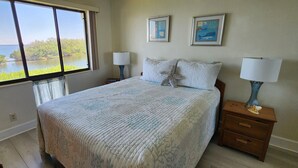 Une chambre avec une grande fenêtre donnant sur un plan d’eau, un lit avec une literie à motifs, deux lampes de chevet et deux tableaux encadrés accrochés au mur.