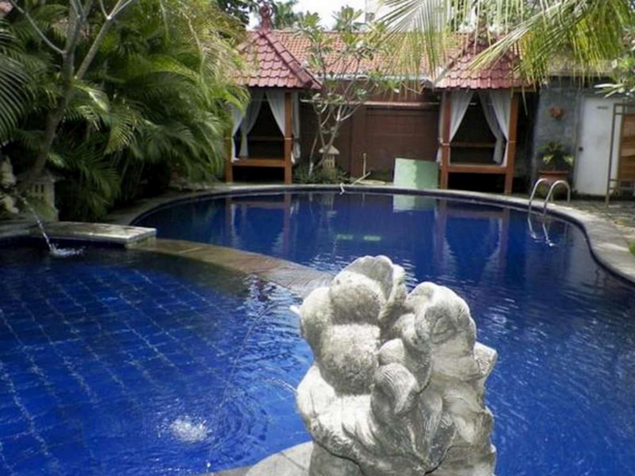 A swimming pool with a stone sculpture in the center, surrounded by tropical plants and a building with a tiled roof in the background.