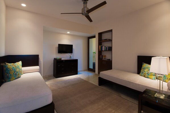 Habitación de hotel con dos camas, televisión, estantería y ventilador de techo.