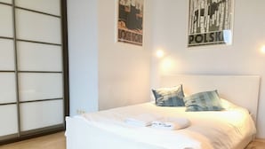A neatly made bed with pillows and a folded towel, a wooden floor, and two posters on the wall. - Apartament balkon-Aleje Jerozolimskie 29 (Warsaw)
