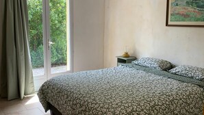 A bedroom with a bed, a nightstand, a painting, and a view of greenery through a window.