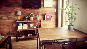 Un interior de madera con un televisor montado en la pared, una mesa de madera con sillas, un estante con objetos y una ventana con una planta.