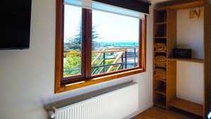 Habitación con un amplio ventanal que da a un balcón con vistas a zonas verdes y un paisaje distante. Debajo de la ventana, hay un radiador y, a la derecha, una estantería de madera.