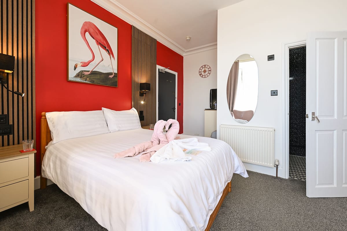 A hotel room with a bed, a nightstand, a mirror, a red wall, and a door.