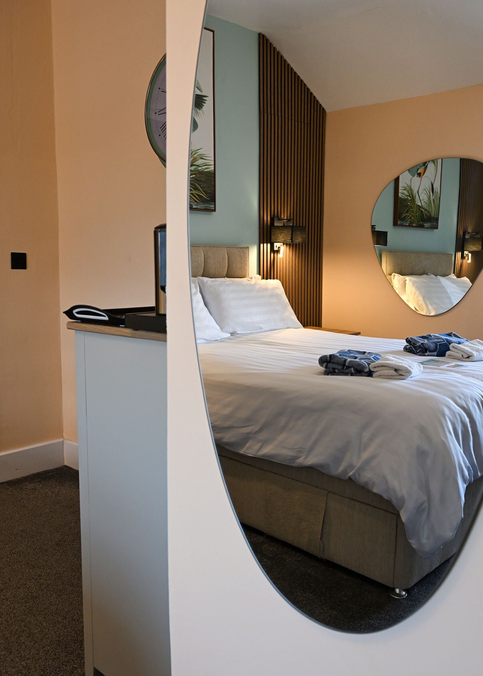 A bedroom with a bed, a desk, a mirror, and a wall-mounted clock.