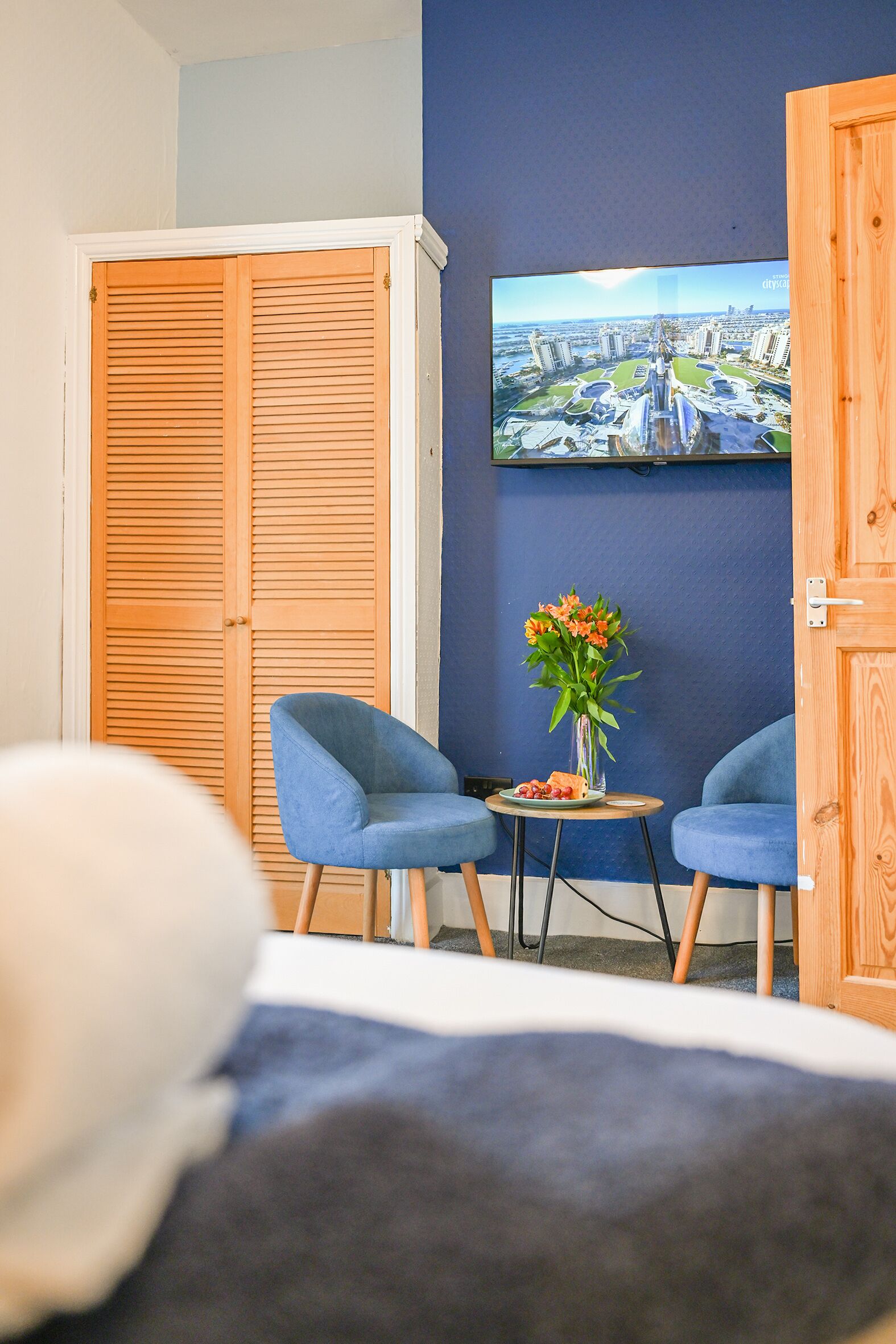 Een hotelkamer met een bed, twee blauwe fauteuils, een bijzettafeltje met een bloemenvaas, en een grote televisie aan de muur.