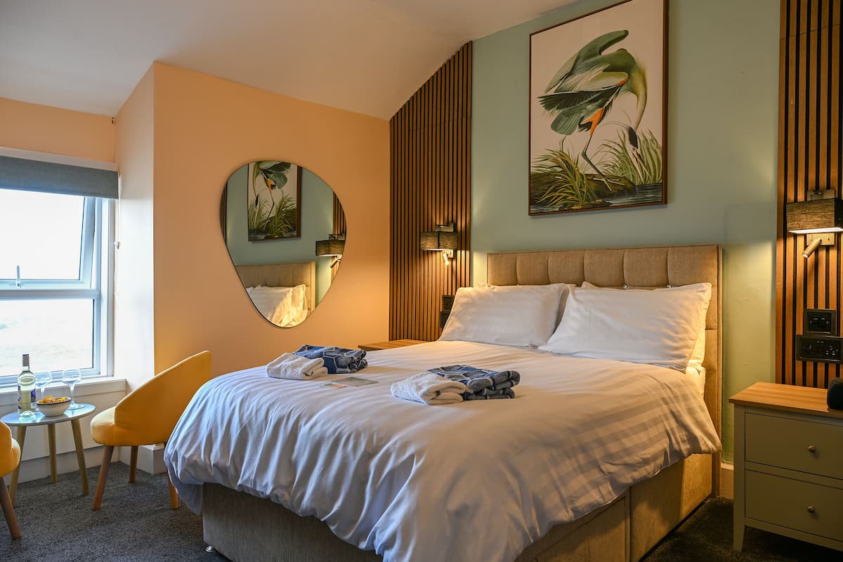A hotel room with a bed, a chair, a bedside table, a mirror, and a window.