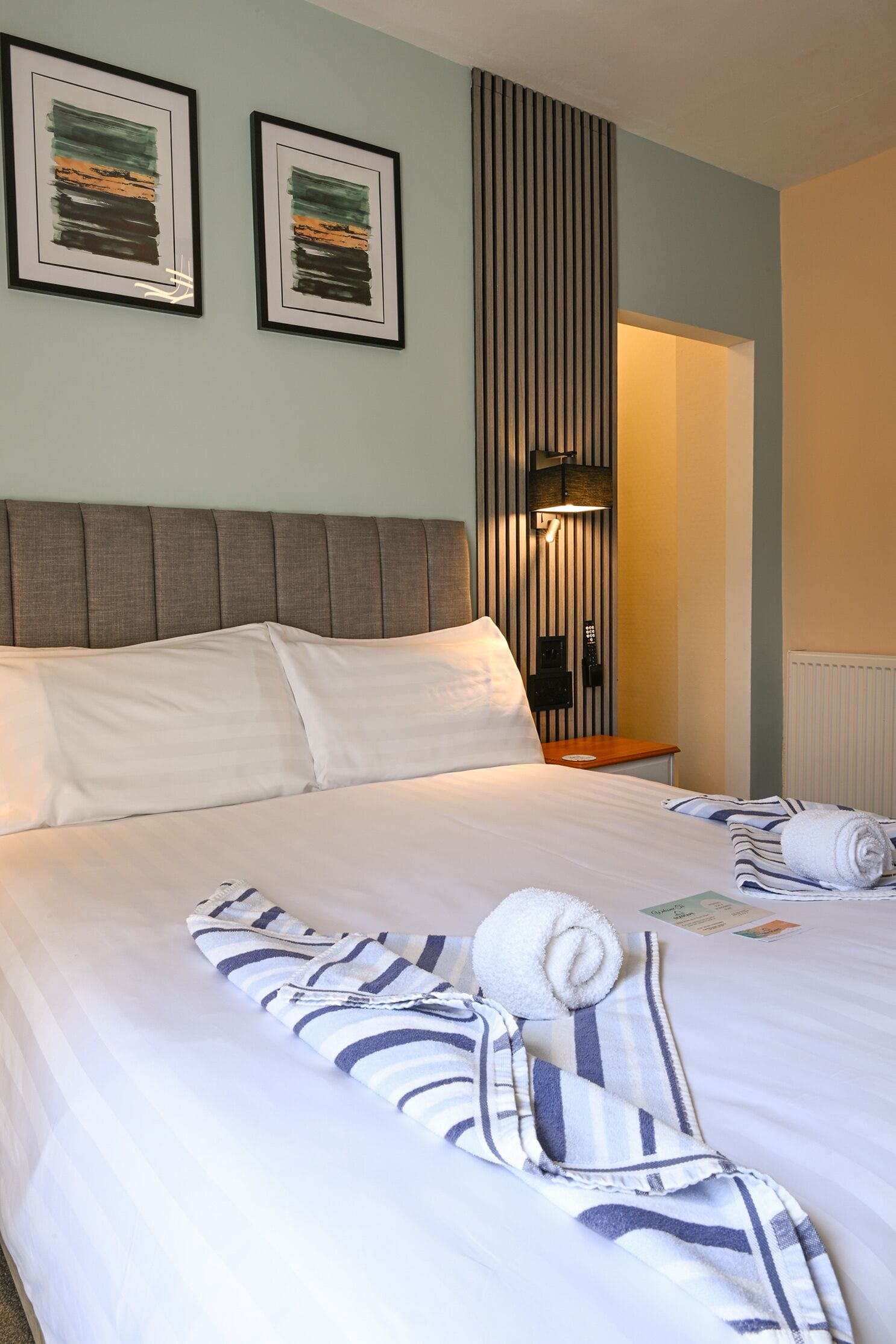 A neatly made bed with pillows, a striped bathrobe, and a rolled towel on the bed. There are two framed artworks on the wall.