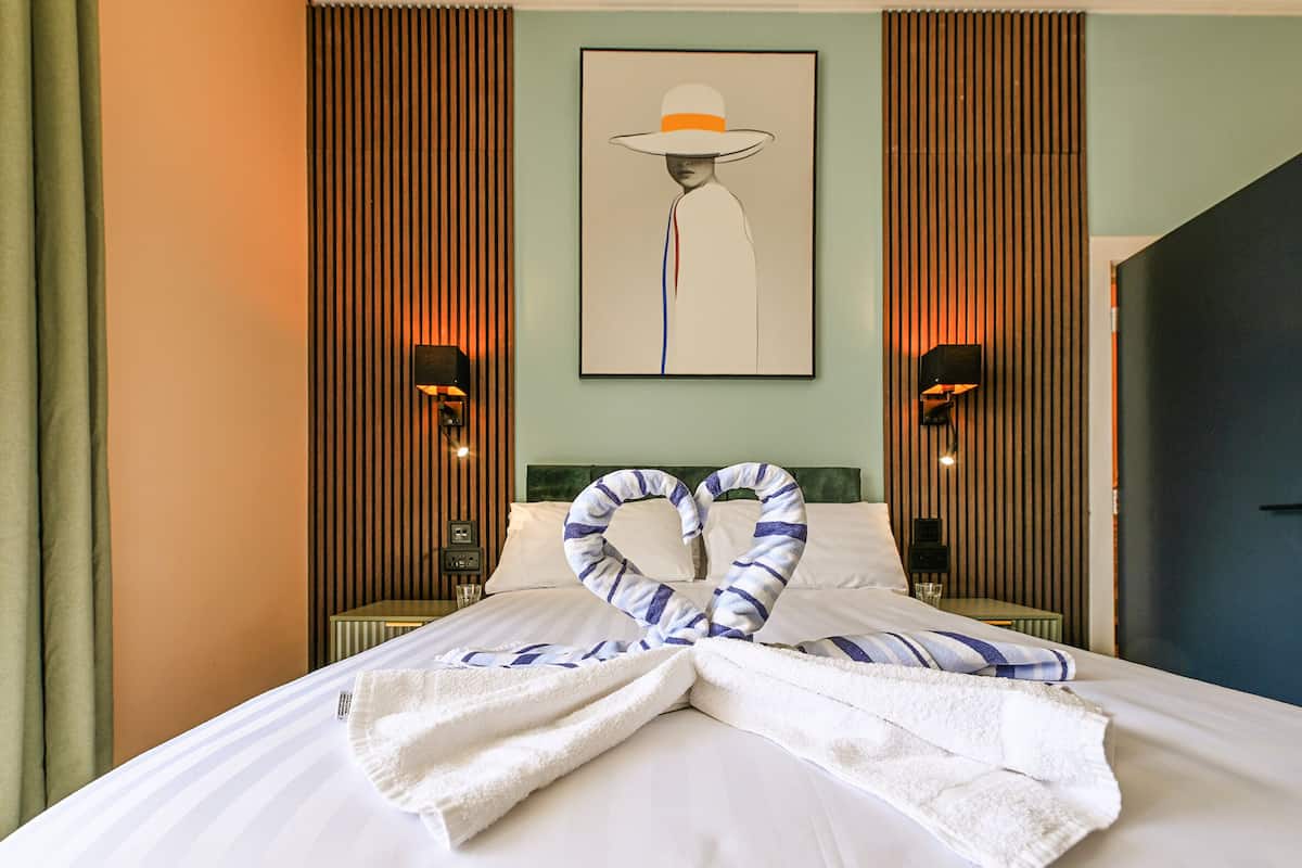 A hotel room with a bed, a heart-shaped towel arrangement, a painting on the wall, and two wall-mounted lamps.