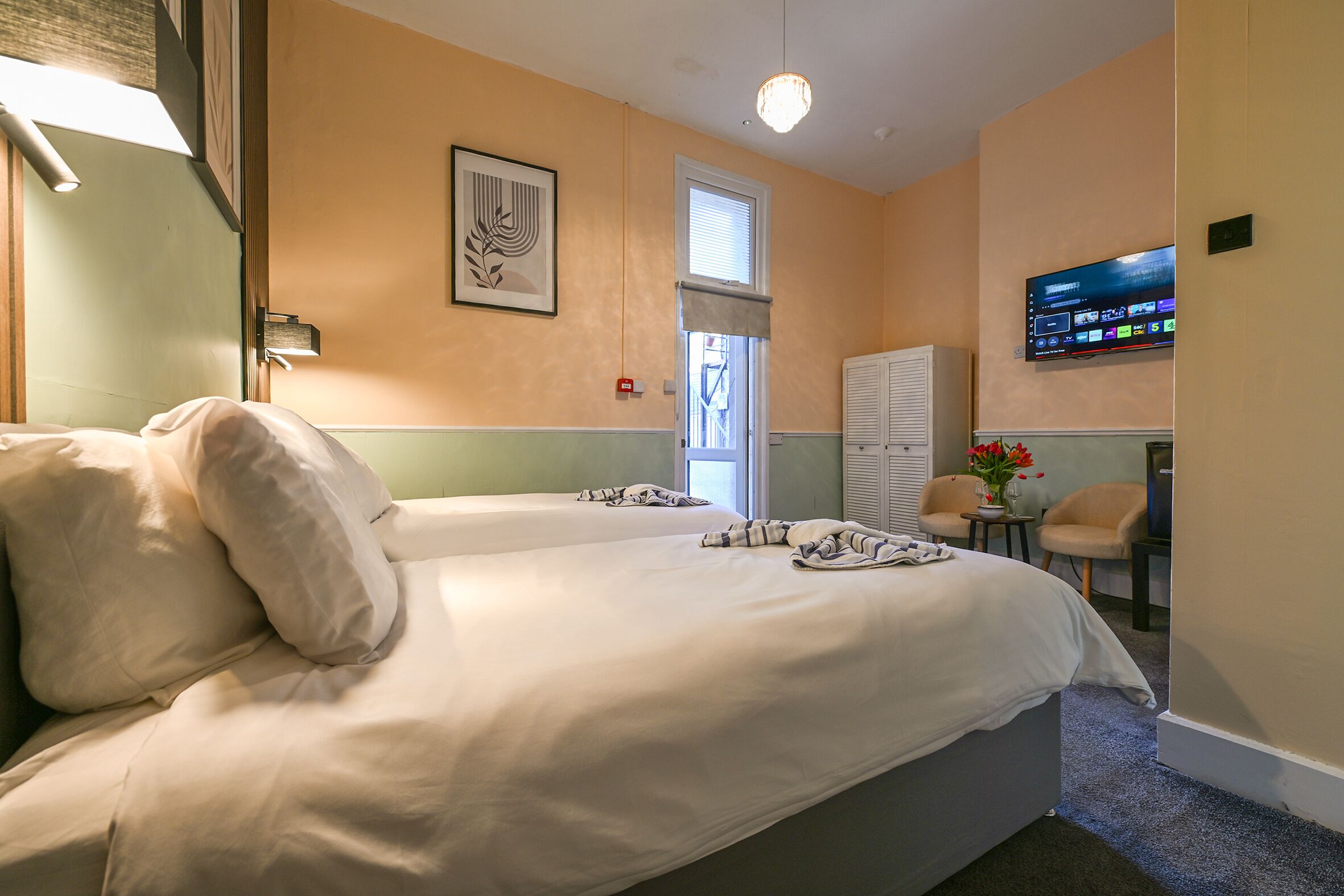 A hotel room with two beds, a television, a window with blinds, and a small table with a vase.