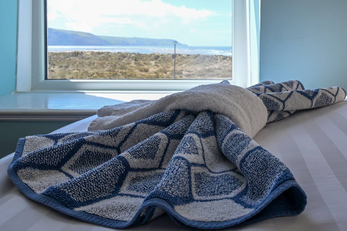 A bed with a blanket and a window with a view of a beach and mountains.