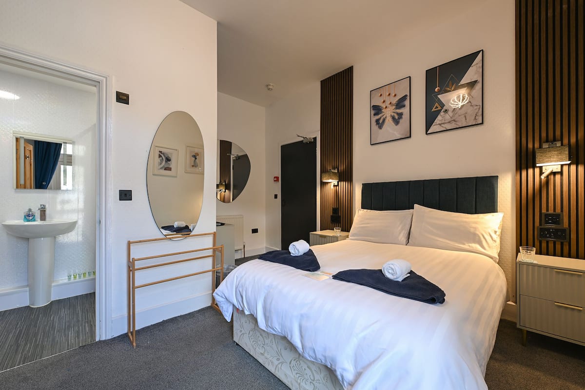 A hotel room with a bed, a bedside table, a nightstand, a mirror, a towel rack, and a wall-mounted light fixture.