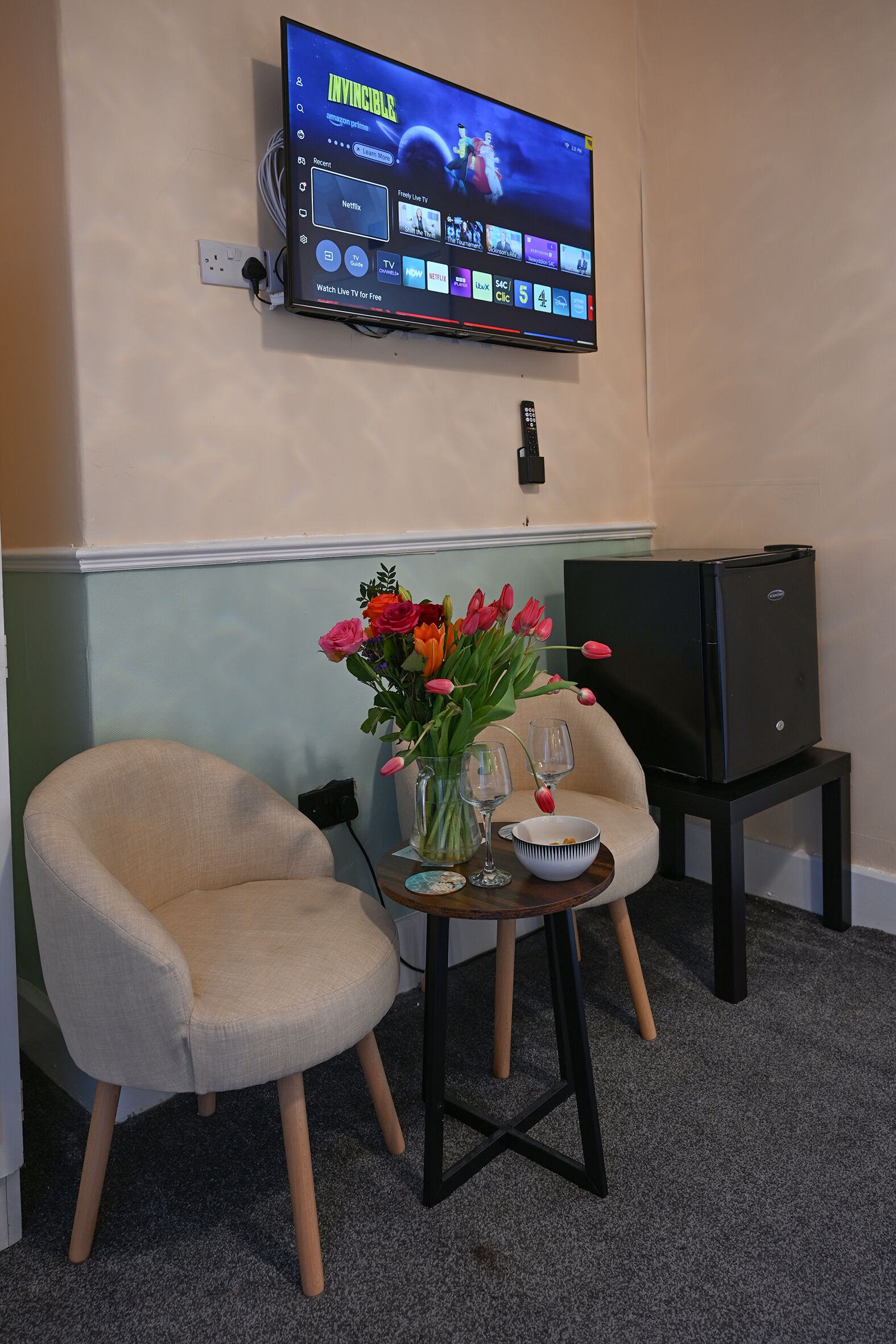 A room with a flat-screen TV mounted on the wall, a beige armchair, a small table with a vase of flowers, a wine glass, and a bowl.