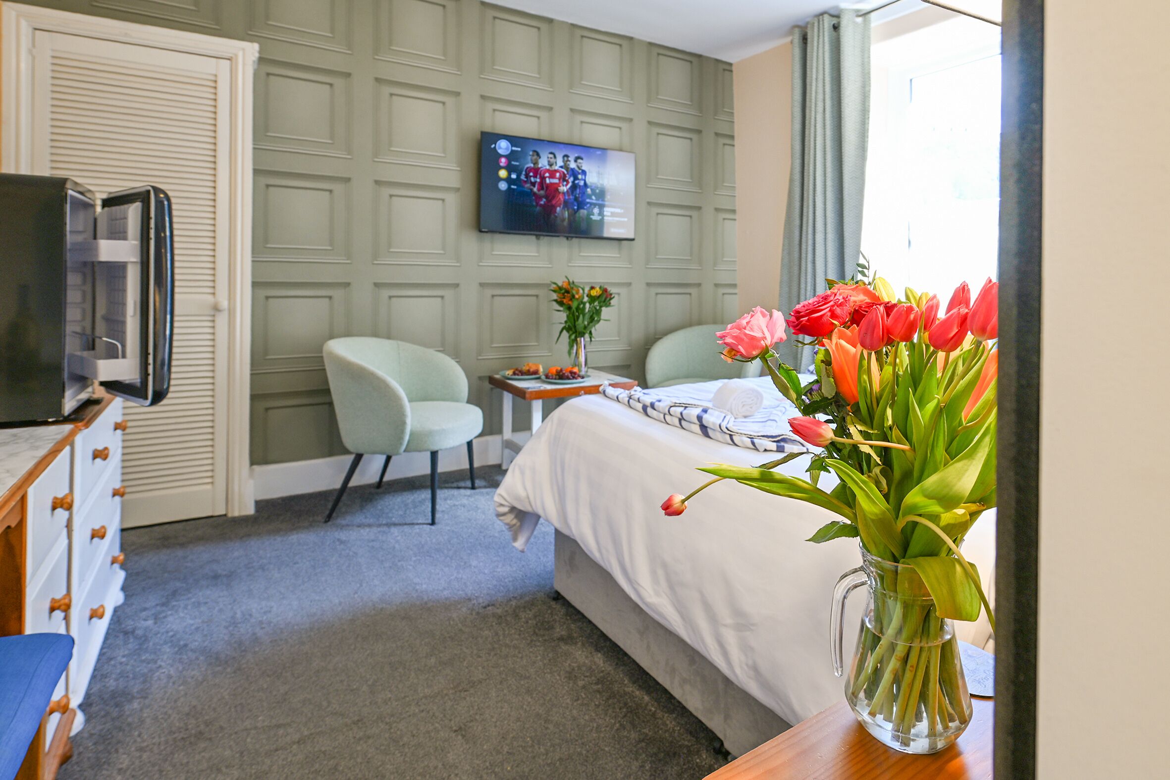 A hotel room with a bed, a TV mounted on the wall, a desk with a vase of flowers, and a chair.