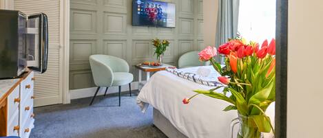 A hotel room with a bed, a TV mounted on the wall, a desk with a vase of flowers, and a chair.