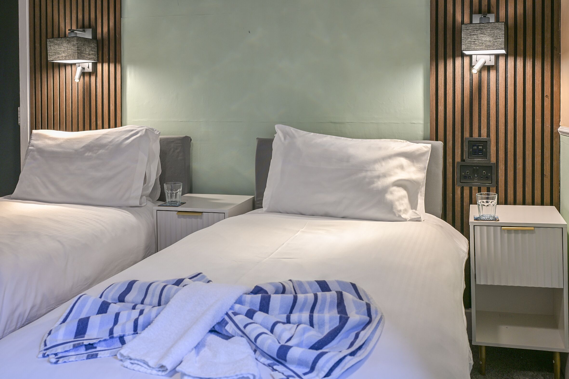 A hotel room with two beds, a nightstand with a lamp, a power outlet, and a wall-mounted control panel.