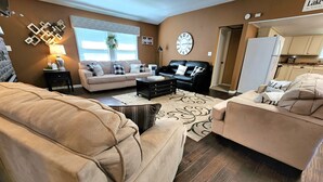 En stue med en beige sofa, en sort lædersofa og et sofabord. Der er et ur på væggen og et vindue med persienner.