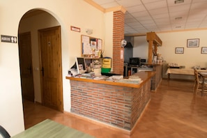 Uma área de recepção com balcão de tijolos, portas de madeira, um mural de avisos e uma sala de jantar com mesa e cadeiras.