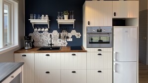 Una cocina moderna con armarios blancos, horno empotrado y refrigerador. Hay dos repisas blancas sobre el fregadero con diversos utensilios de cocina.