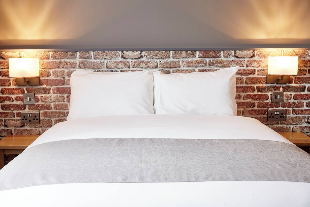 A bed with white linens, a grey throw, and a brick wall with two wall-mounted lamps.