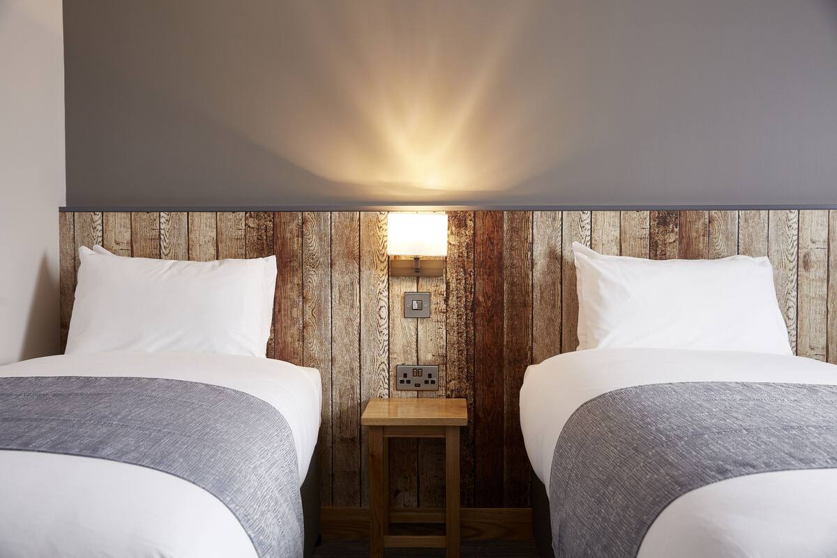 A room with two beds, a wooden wall, a small table with a lamp, and a power outlet.
