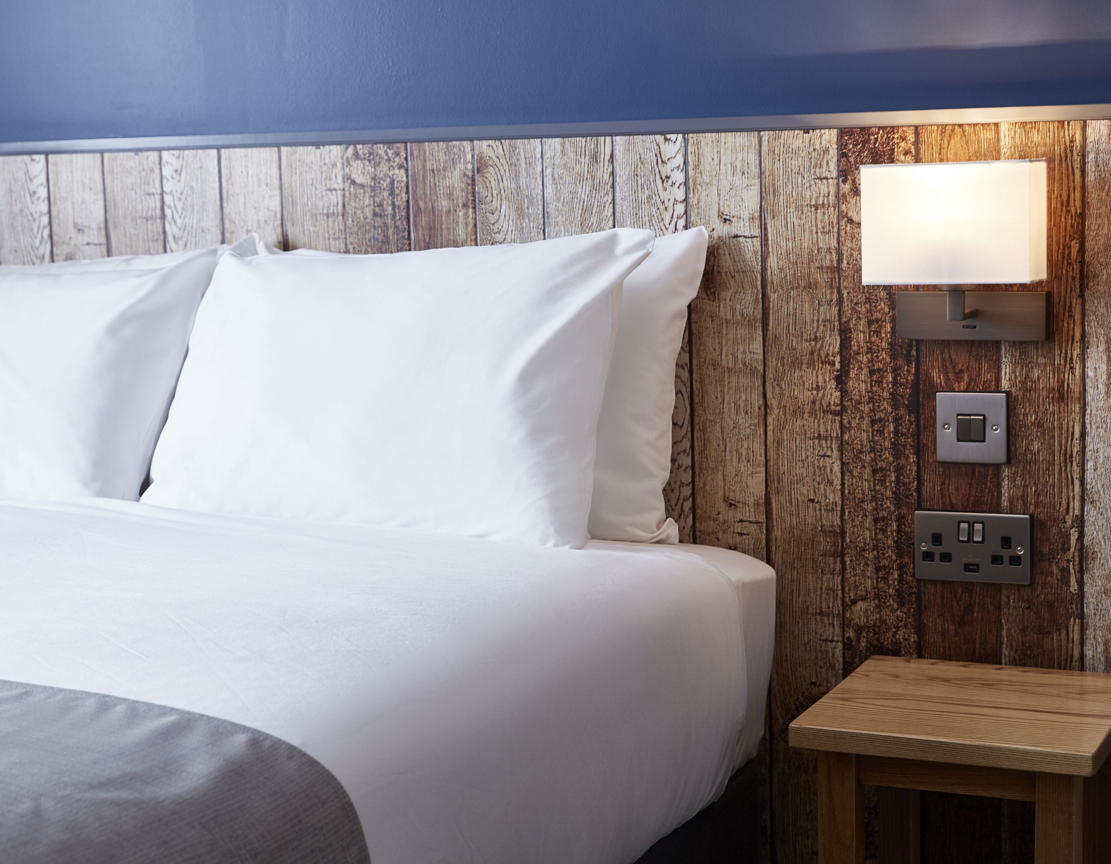 Un lit bien fait, avec du linge de lit blanc, dans une pièce dont un mur est recouvert de lambris en bois et qui est équipée d’un luminaire fixé au mur.