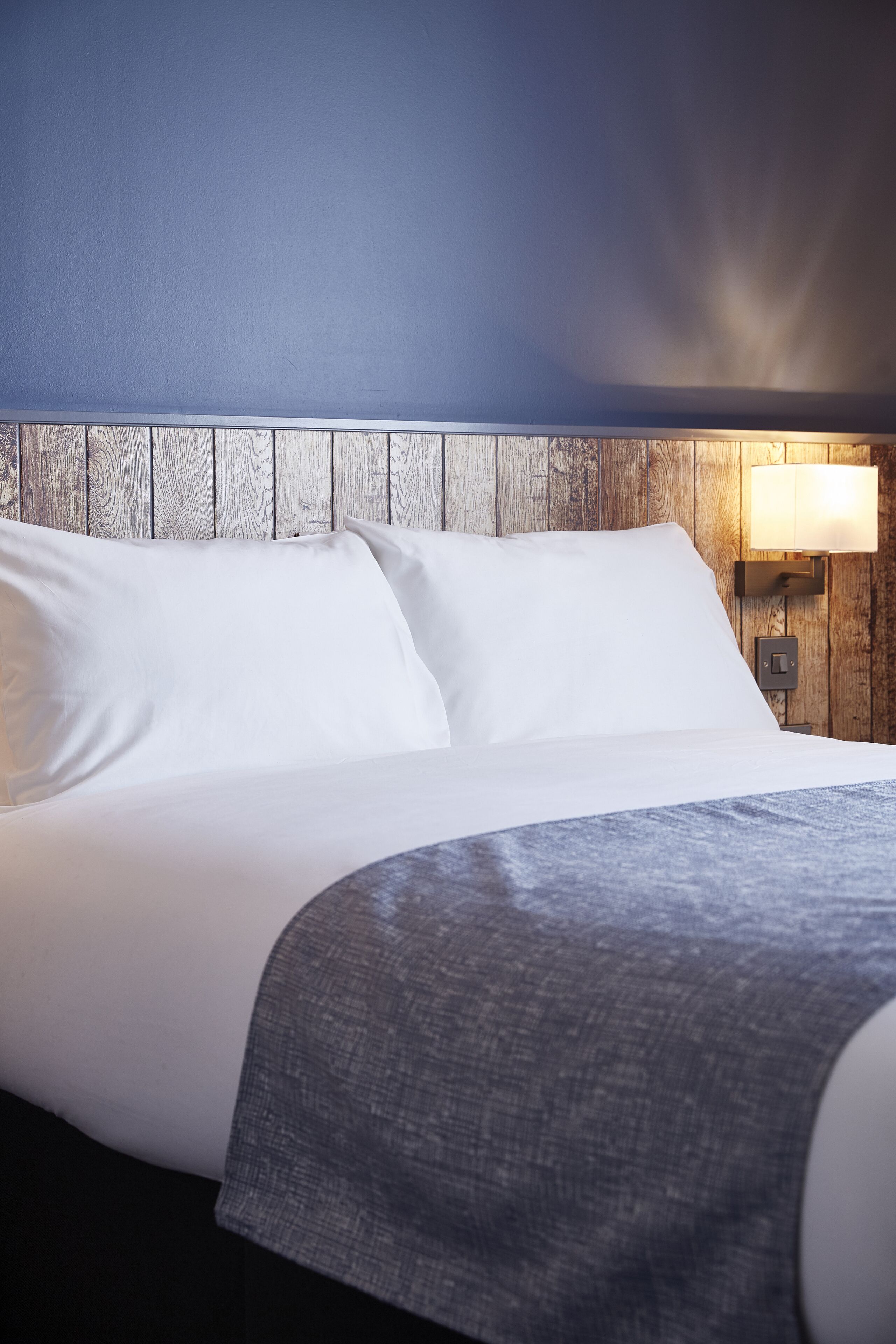 Ein ordentlich bezogenes Bett mit weißen Laken und einer grauen Tagesdecke, vor einem Holzkopfende und einer Wand mit einer Leuchte.