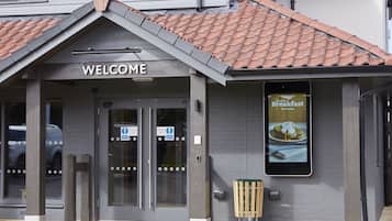 A building entrance with a 'WELCOME' sign, automatic doors, a 'Breakfast' sign, and a trash bin.