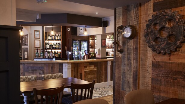 A bar area with a wooden counter, shelves with bottles, a clock, and a gear decoration on the wall. - The Churchill By Greene King Inns (Swindon)