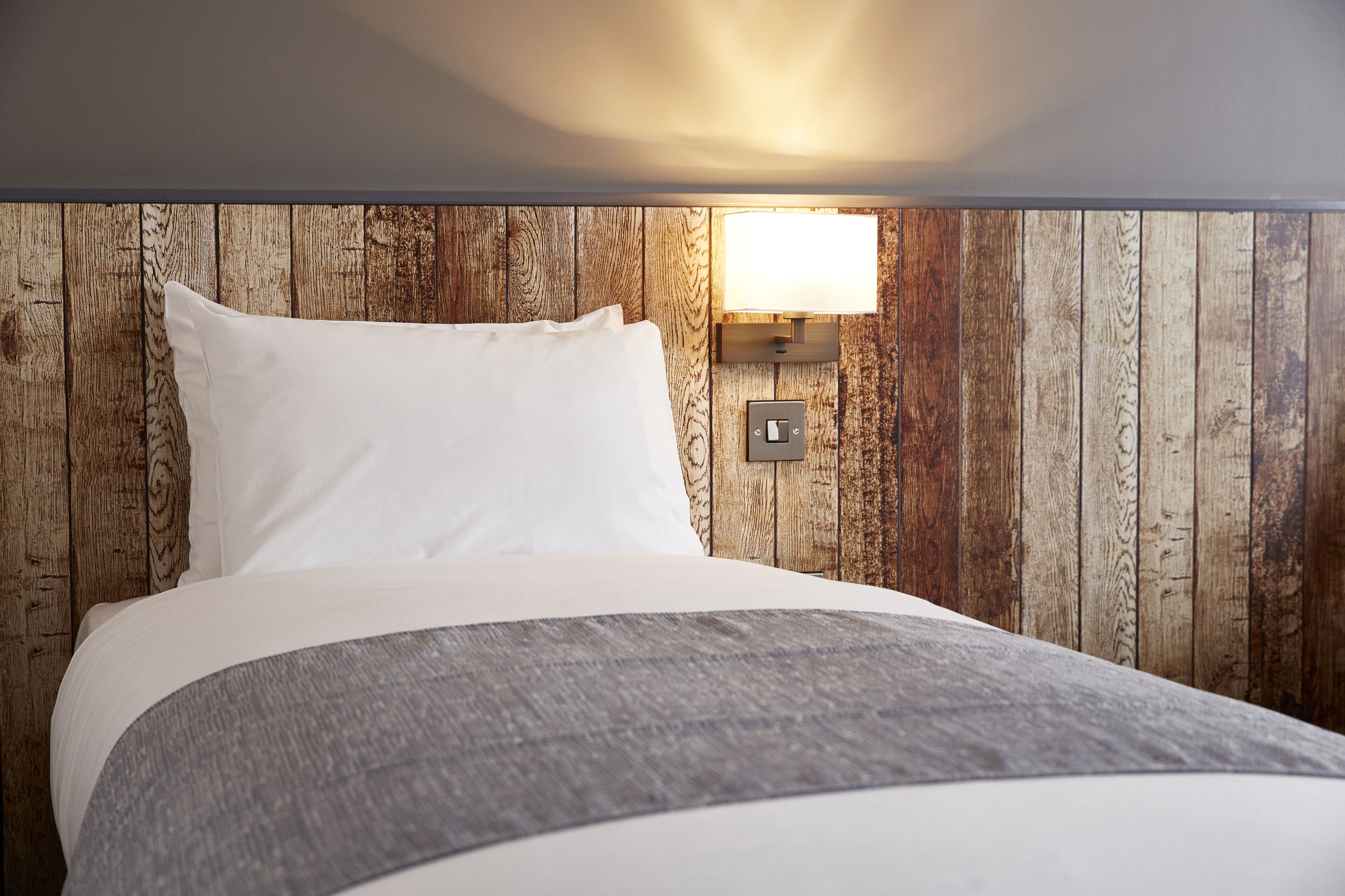 Un lit avec du linge de lit blanc et un plaid gris, un lambris en bois et une lampe fixée au mur.