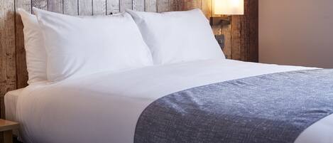 A neatly made bed with white linens and a gray throw blanket, a wooden headboard, and a wall-mounted nightstand with a lamp.