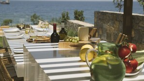 A table set for a meal with a view of the sea, including plates, glasses, a bottle of wine, cheese, grapes, and tomatoes.